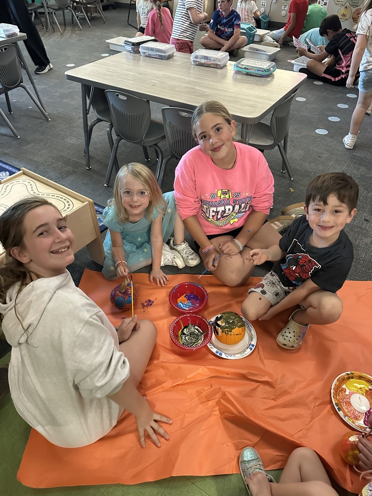 students smiling at a camera painting pumpkins on the floor of a classroom