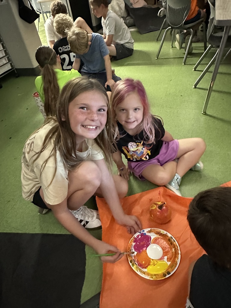 two students smiling at the camera painting a pumpkin on the floor