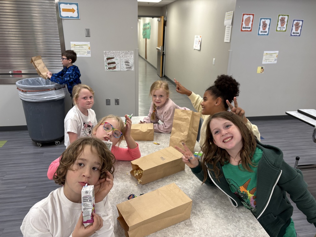 Group of elementary students sitting along a cafeteria table during snack time, making peace signs and smiling for the camera.