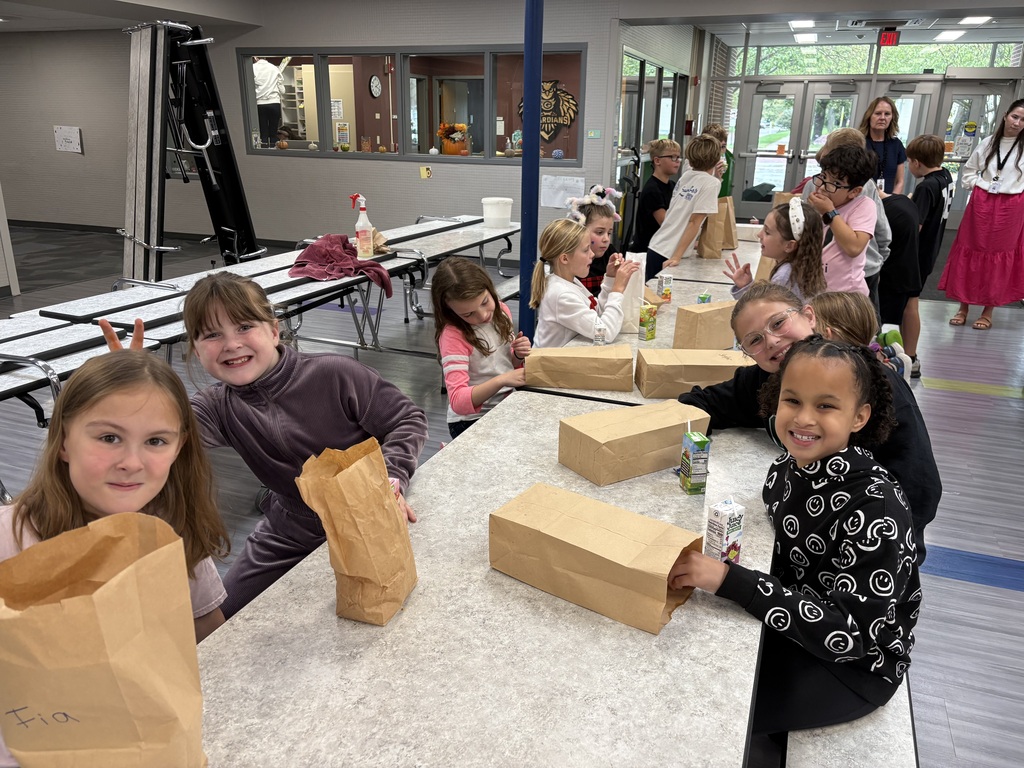 Large group of young students sitting along cafeteria tables with brown paper sacks and juice boxes, smiling together in a brightly lit hallway area.