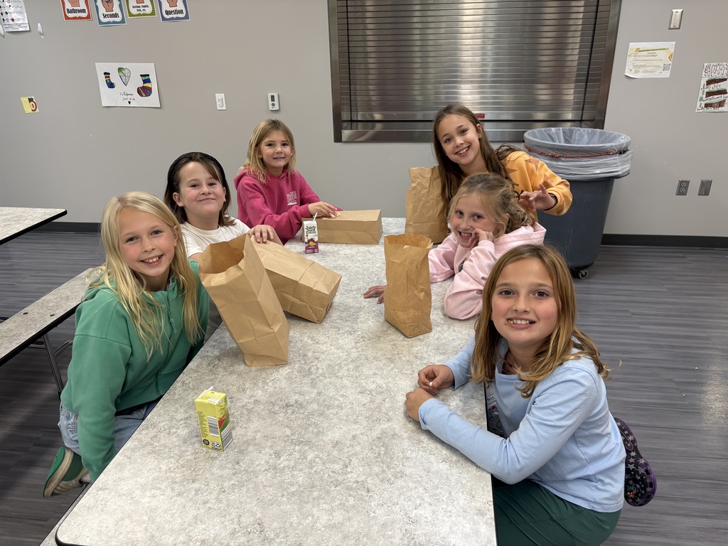 Group of six young girls sitting around a lunch table in a school cafeteria, smiling together with paper lunch sacks and juice boxes.