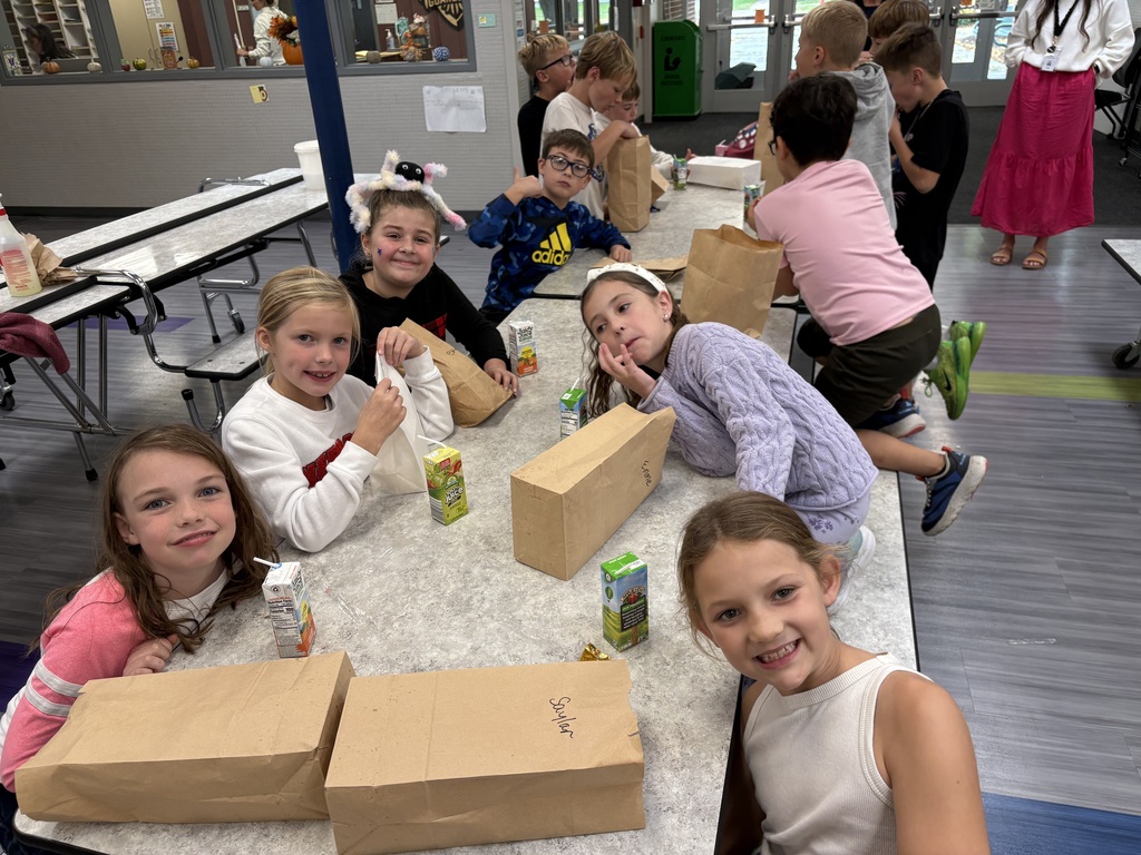 Group of elementary school girls sitting around a cafeteria table during lunch, smiling together next to brown paper bags and juice boxes.