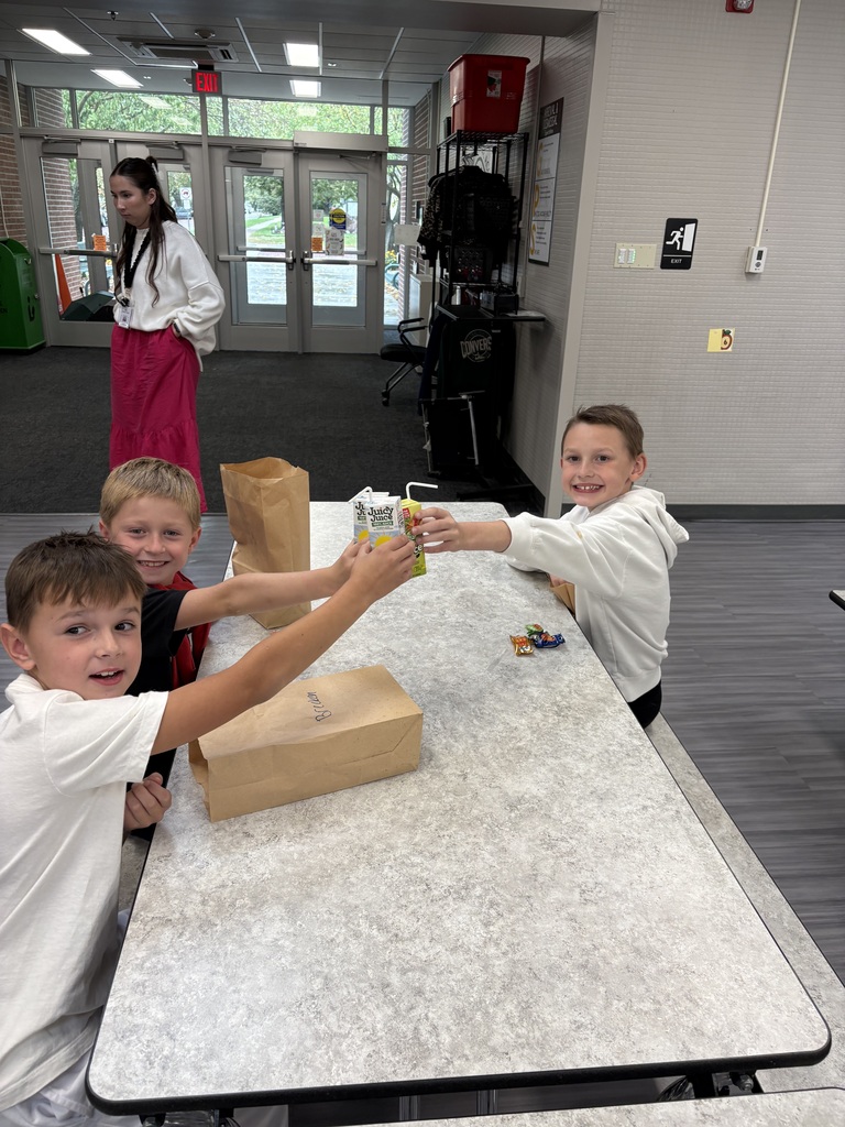 Three young boys sitting at a school cafeteria table, smiling and clinking their juice boxes together in a toast over paper lunch bags.