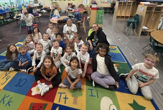Elementary students sit cross-legged on a colorful alphabet rug in a classroom, posing together for a group photo with two teachers behind them.