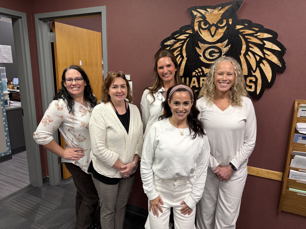 Five female school staff members smiling together for a group portrait in an office in front of a wooden owl mascot sign.