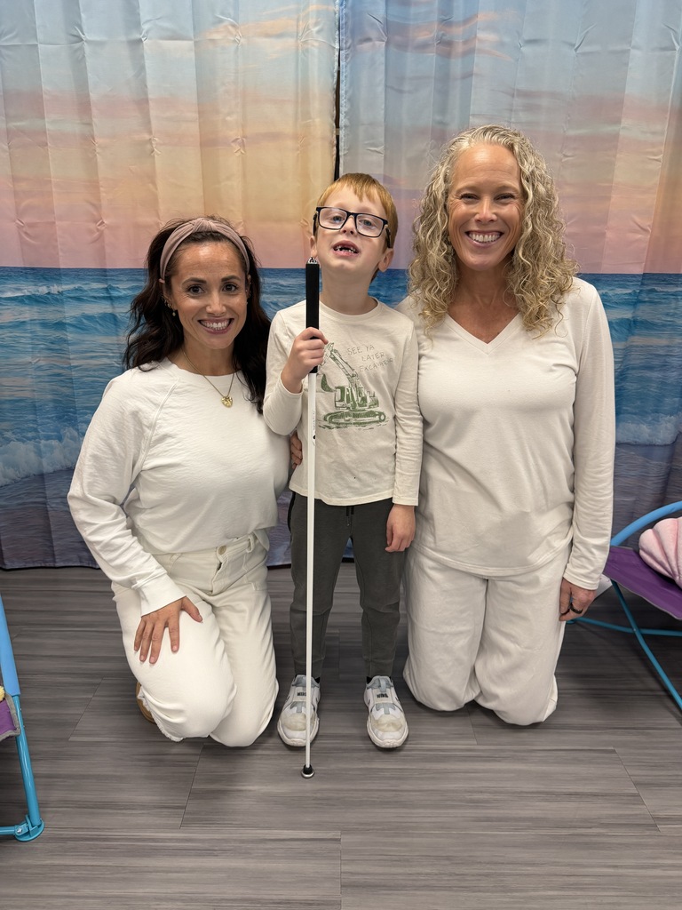 A young boy holding a white cane stands between two smiling women in front of a beach-themed photo backdrop.