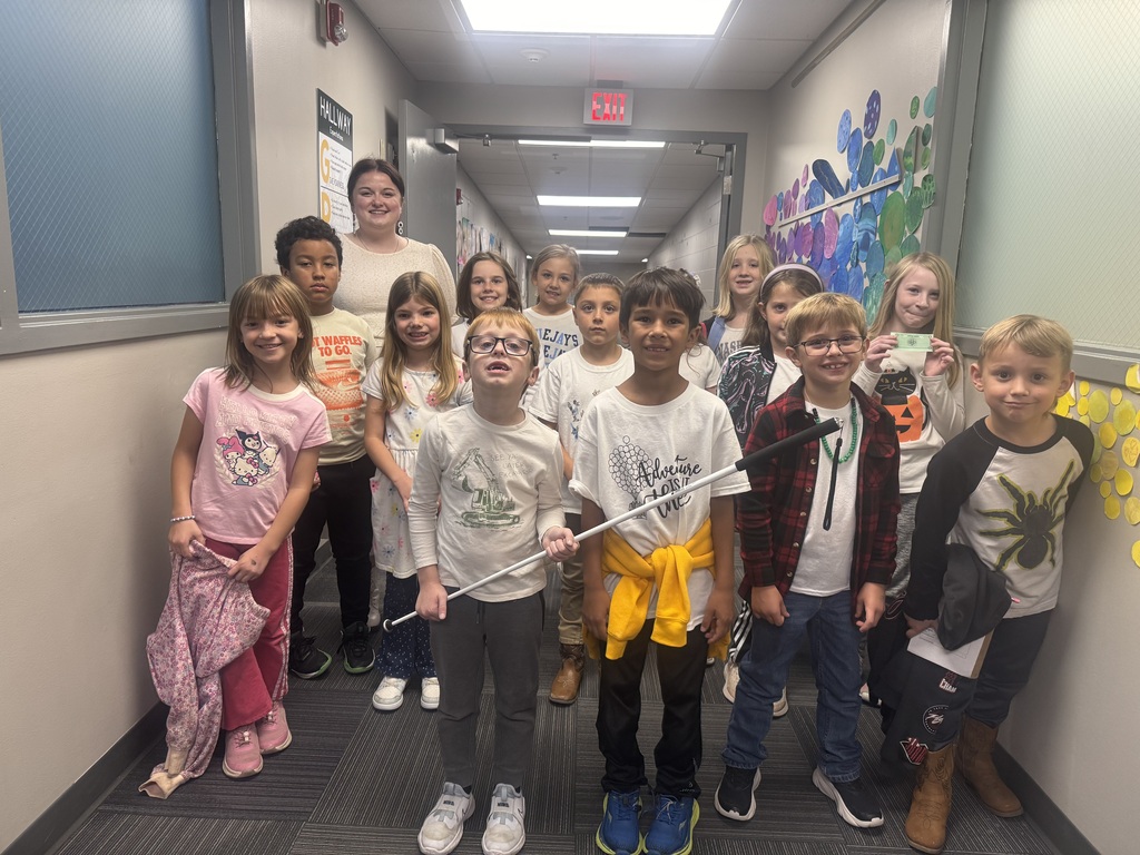 A group of elementary school students and a teacher gather in a carpeted school hallway for a group photo, with one boy holding a white cane.