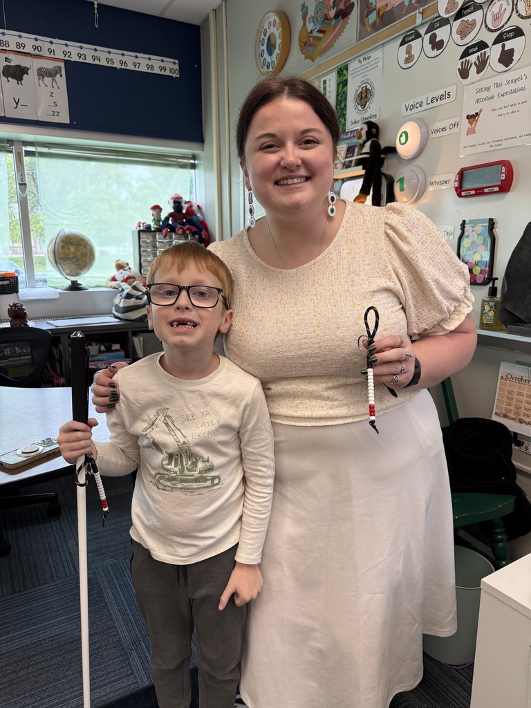 A young boy holding a white cane poses indoors next to a smiling female teacher who holds a beaded keychain.