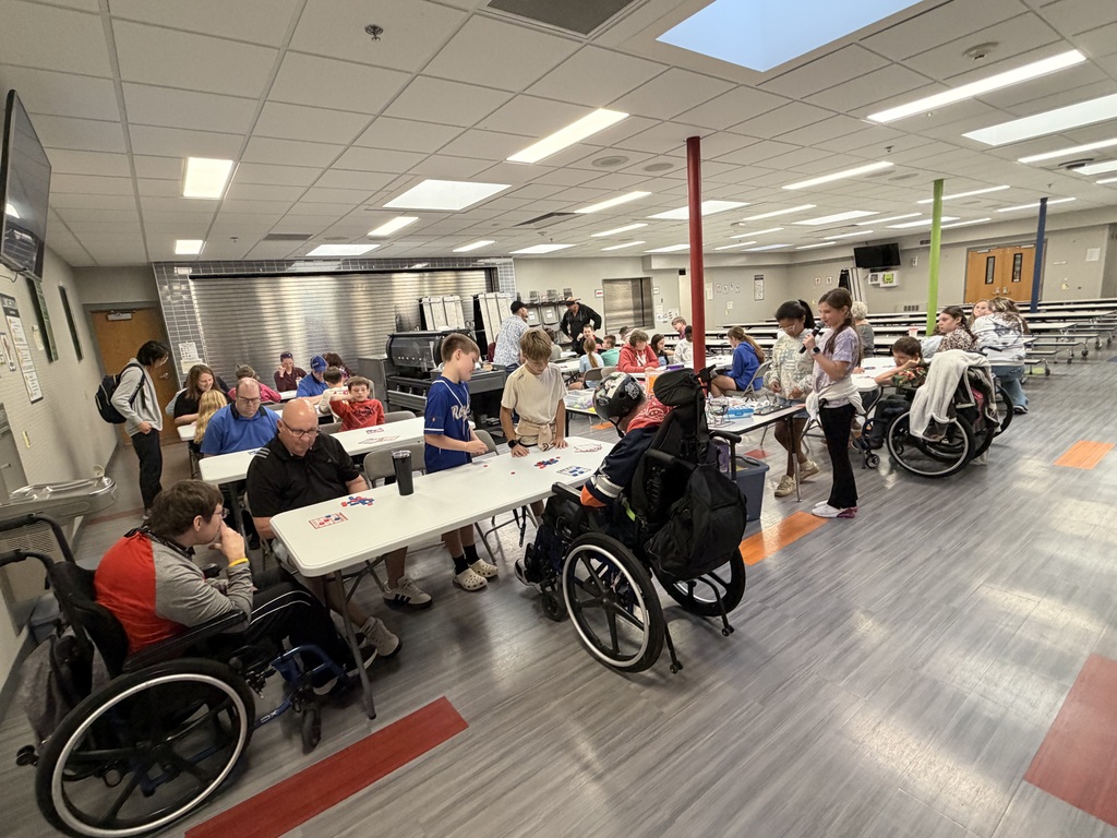 Wide view of a school cafeteria filled with participants sitting at tables playing bingo, with students leading the activity from a table with prizes.