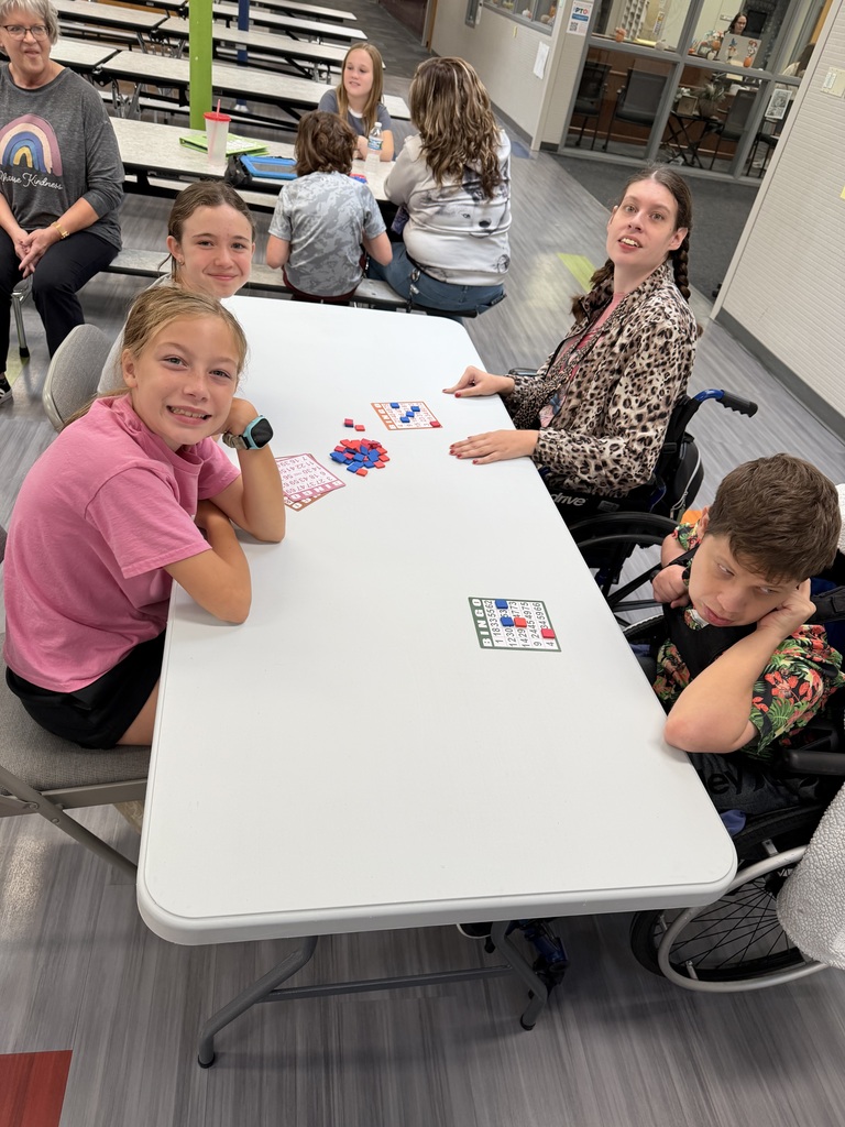 Four participants sitting at a white table in a cafeteria, smiling as they play a game of bingo with cards and red and blue markers.