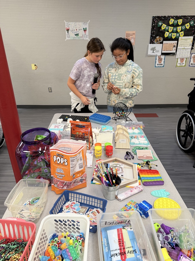 Two elementary school girls standing behind a prize table filled with toys and candy, holding a microphone and small prize while calling out numbers for a game.