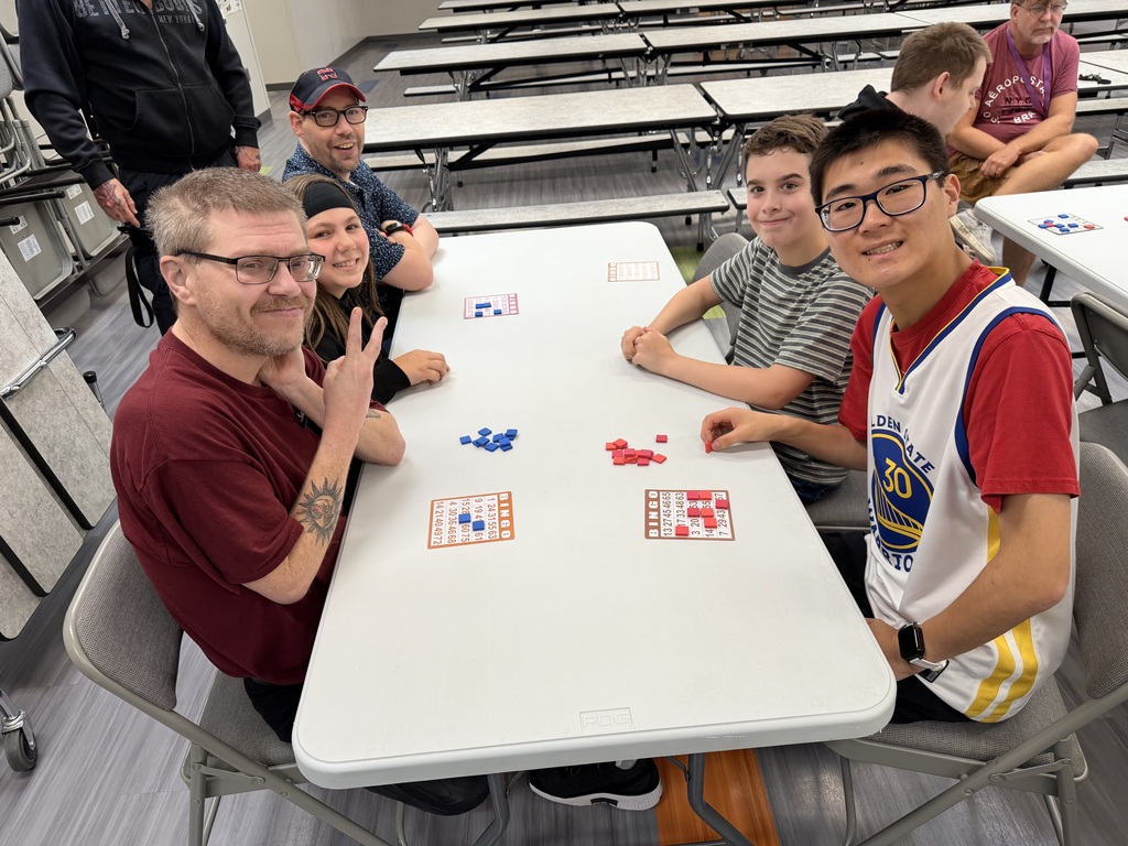 Four participants sitting around a table in a school cafeteria, smiling and holding up a peace sign while playing bingo.