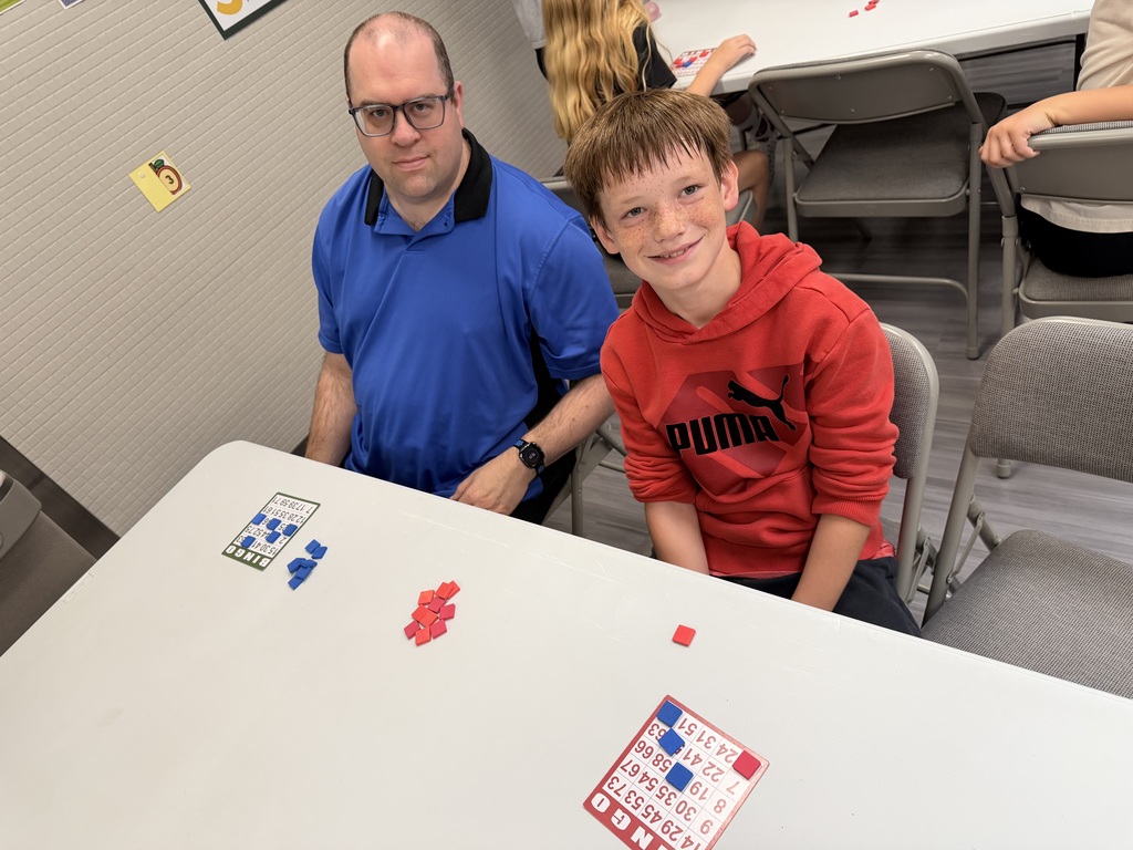 An adult man in a blue shirt and a young boy in a red Puma hoodie sitting together at a table with bingo cards spread out in front of them.