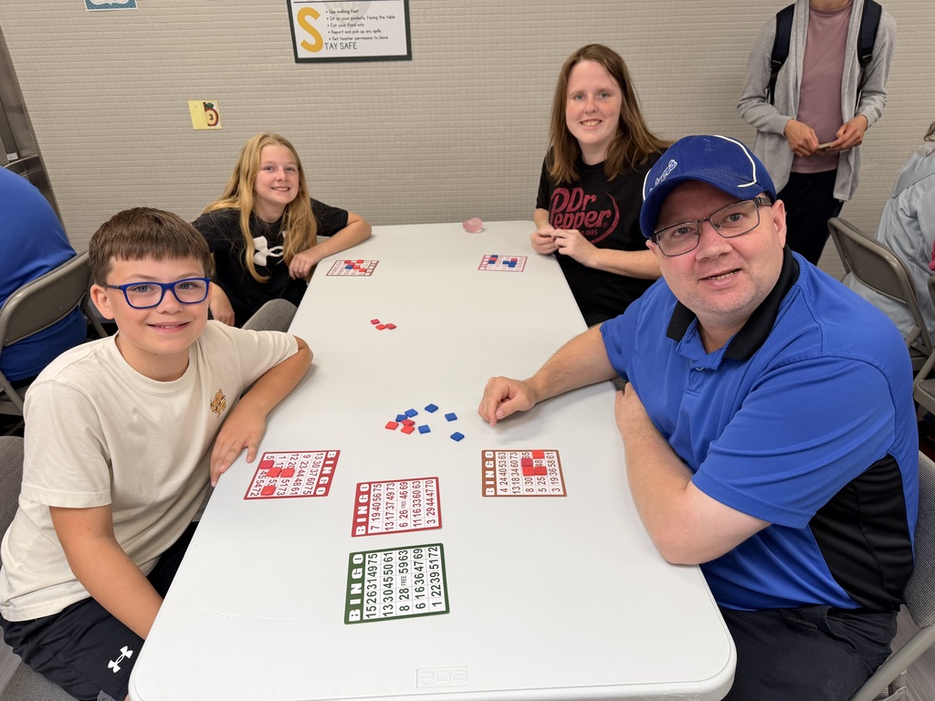 A boy and a man sitting together at a white table in a cafeteria, smiling at the camera with bingo cards set out before them.