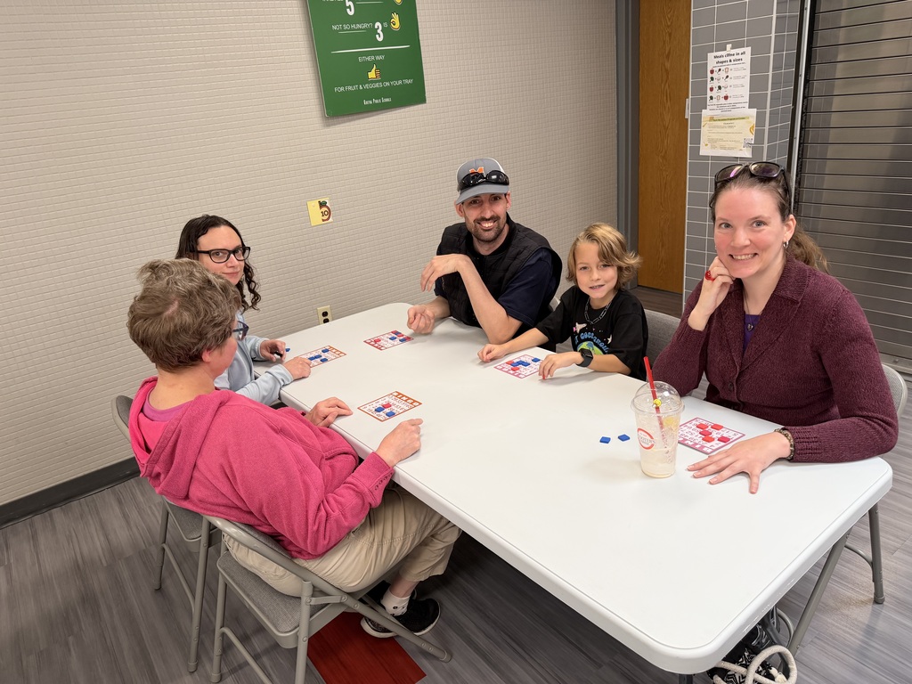 Family group sitting at a white cafeteria table, smiling together as they play a game of bingo with colored markers.