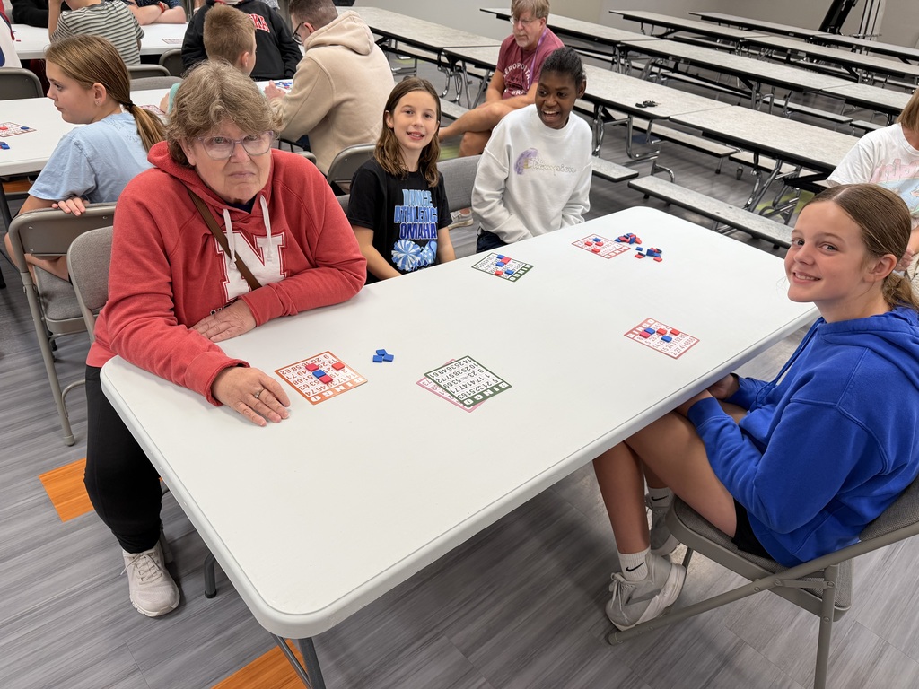 Group of five adults and children sitting around a long folding table in a school cafeteria, smiling while playing bingo together.