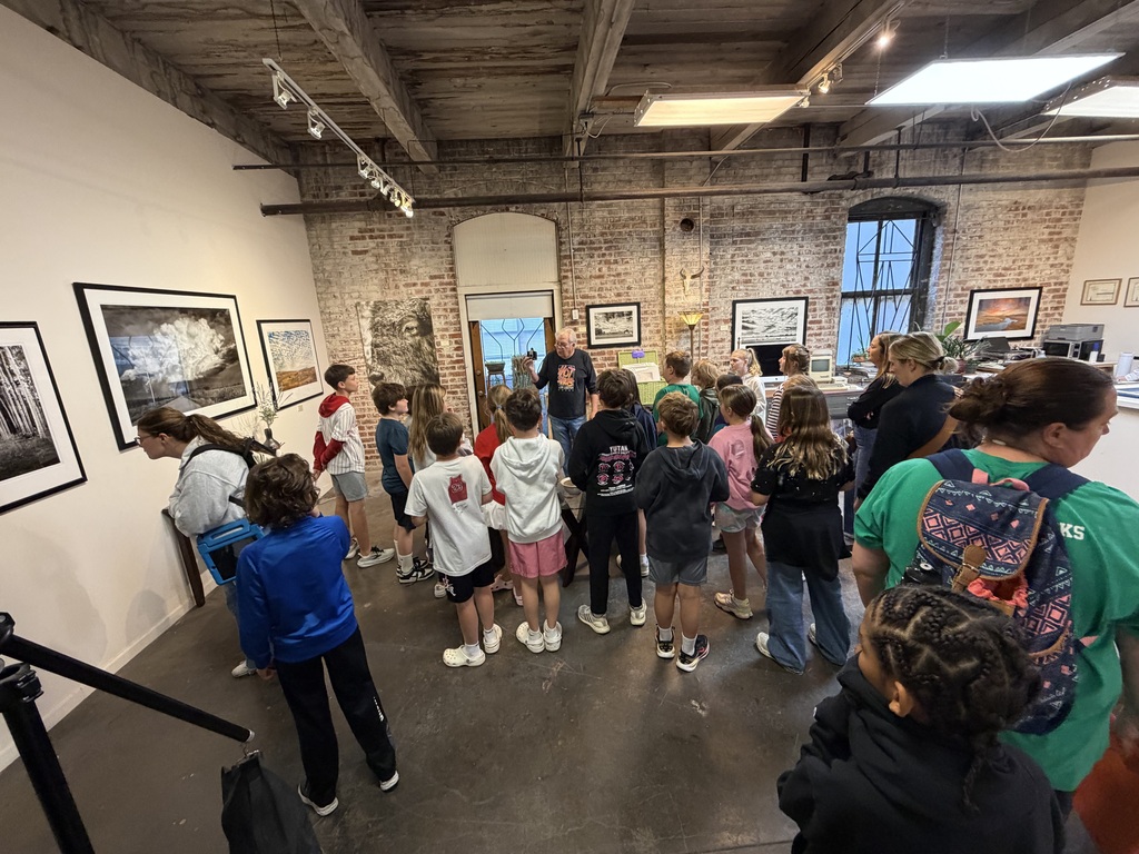 Group of elementary students gather inside an art gallery with brick walls to view framed black and white photographs.