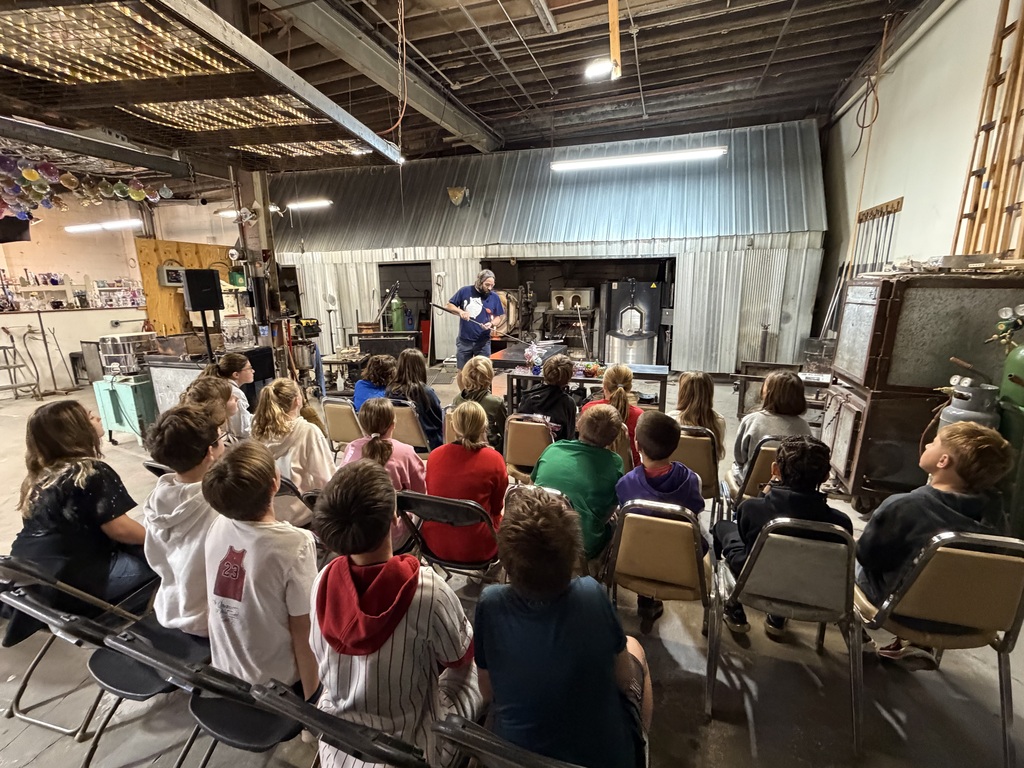 Students seated in rows watch a glassblowing demonstration inside an industrial workshop with active kilns in the background.