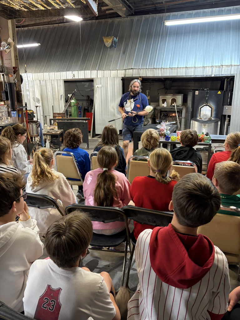 A glassblower demonstrates working with molten glass on a blowpipe for a audience of seated students inside a workshop.