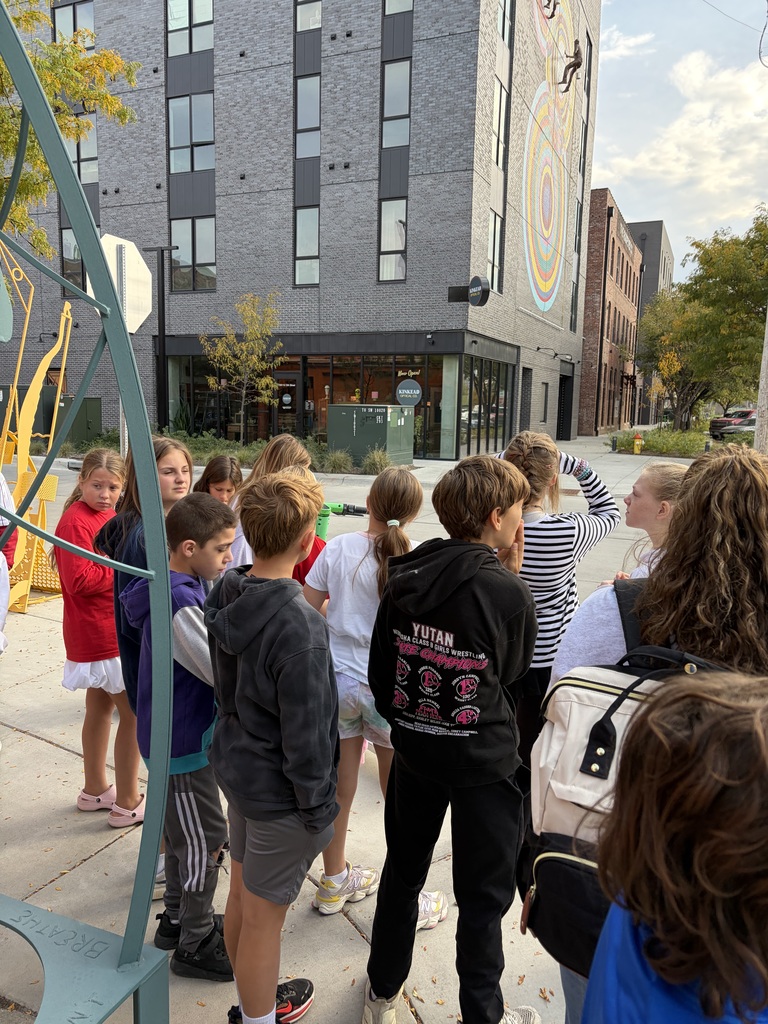 Students stand on a city sidewalk looking up at a tall brick building adorned with a large mural.