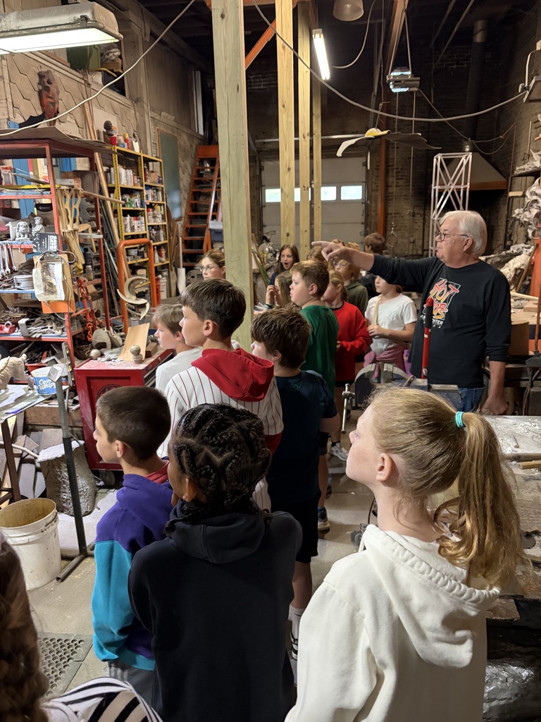 An instructor gestures and speaks to a group of attentive students touring an artist workshop and sculpture studio.