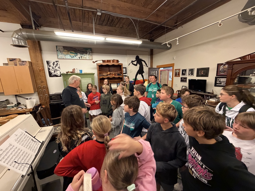 Group of elementary school students surrounding an instructor inside a music room filled with instruments and art decor.
