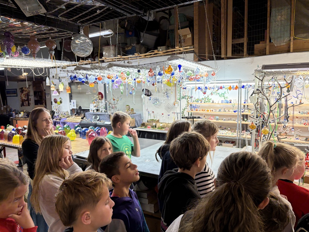 A group of young students gather indoors to view colorful blown glass ornaments and artwork on display shelves.