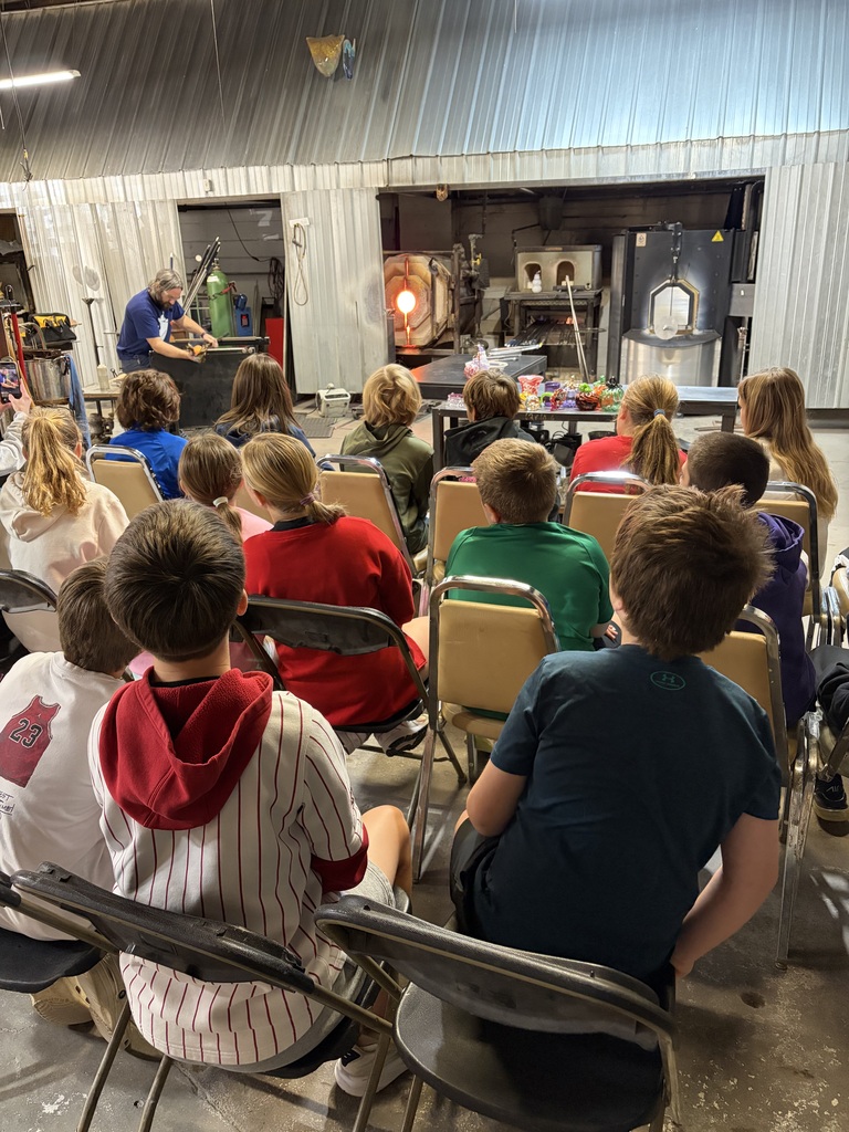 Seated students observe a glassblower working near a glowing hot furnace during a glass studio demonstration.