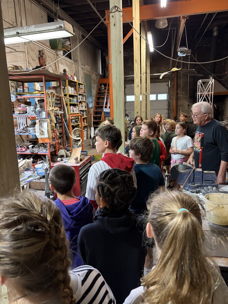 Students gather in a sculpture studio as an instructor speaks near shelves of tools and art equipment.