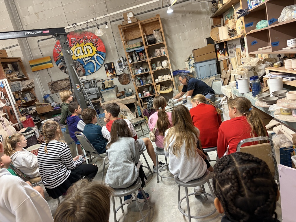 Students sit on stools watching a potter shape clay on a spinning pottery wheel in a ceramics studio.
