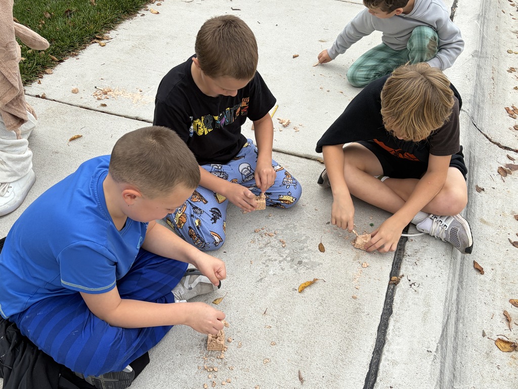 Three young boys sitting on a sidewalk using wooden sticks to excavate fossils from sand blocks during an outdoor lesson.