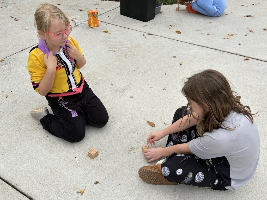 Two elementary students sitting on a concrete walkway, using wooden tools to dig into small clay blocks during a hands-on activity.