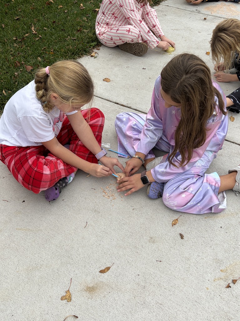 Two girls sitting cross-legged on a sidewalk, collaborating to dig out a fossil block using small wooden sticks.