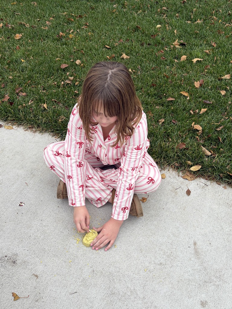 Girl in pink striped pajamas sitting cross-legged on a sidewalk while using a small tool to excavate a yellow fossil block.