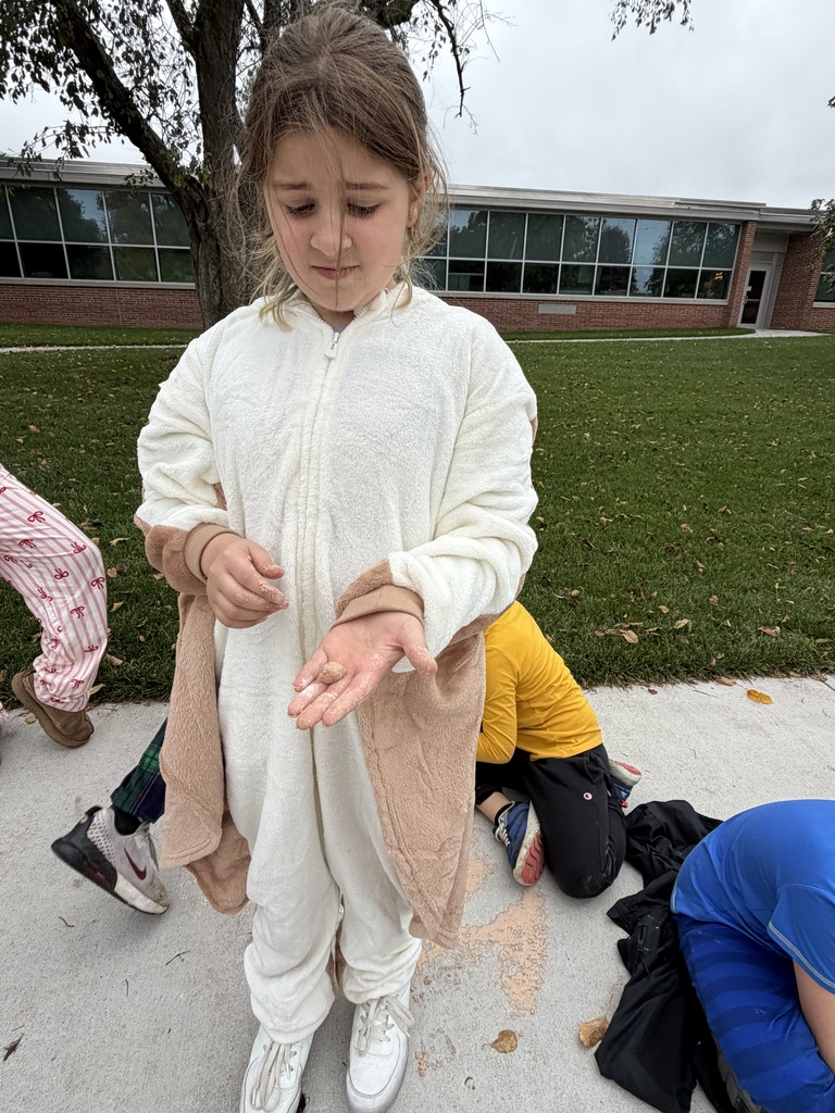 Young girl in a plush hooded onesie standing on a sidewalk, displaying a small fossil specimen in her open palm.
