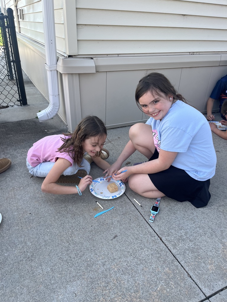 Two elementary school girls laughing while kneeling on a concrete patio to dissect a small fossil excavation block on a paper plate.