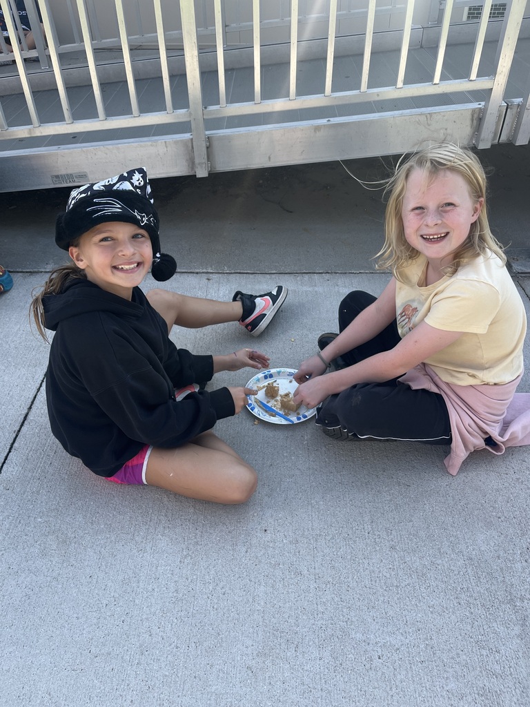 Two young girls sitting cross-legged on a sidewalk, smiling at the camera while excavating a small block on a paper plate.