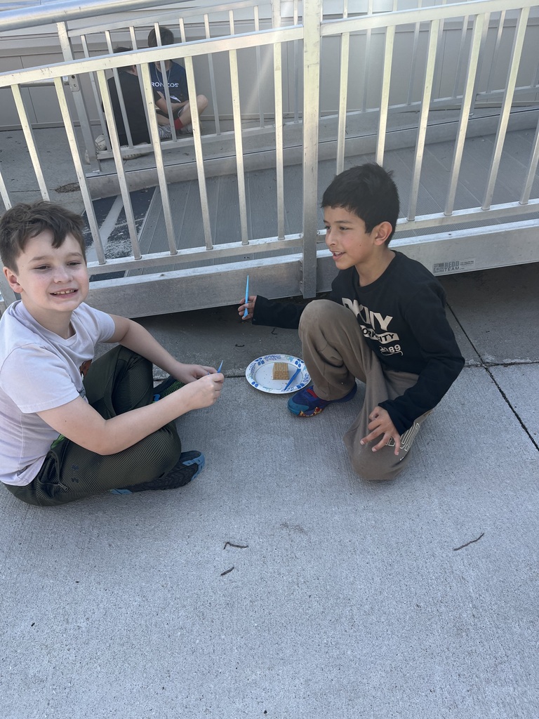 Two boys sitting outdoors on the sidewalk in front of a metal railing, holding plastic tools around a paper plate containing a dig block.