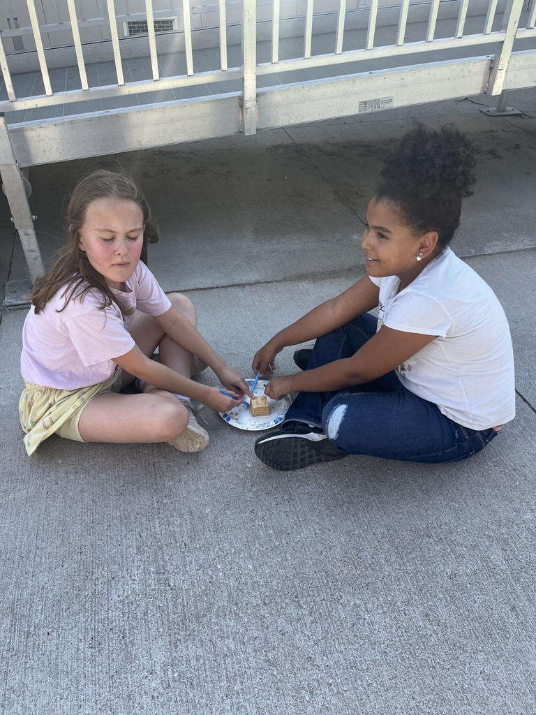 Two elementary school girls sitting opposite each other on a concrete patio, using small plastic tools to work on a dig block together.