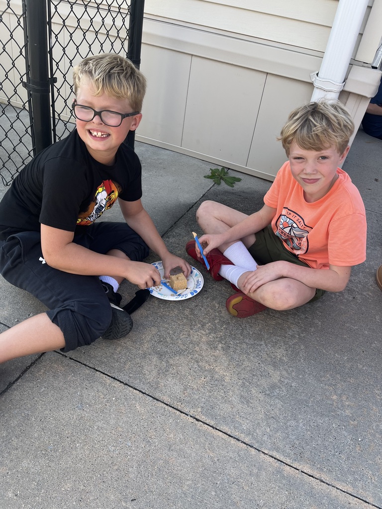 Two boys sitting cross-legged on a concrete ground, using blue plastic tools to chip away at a sand block on a paper plate.