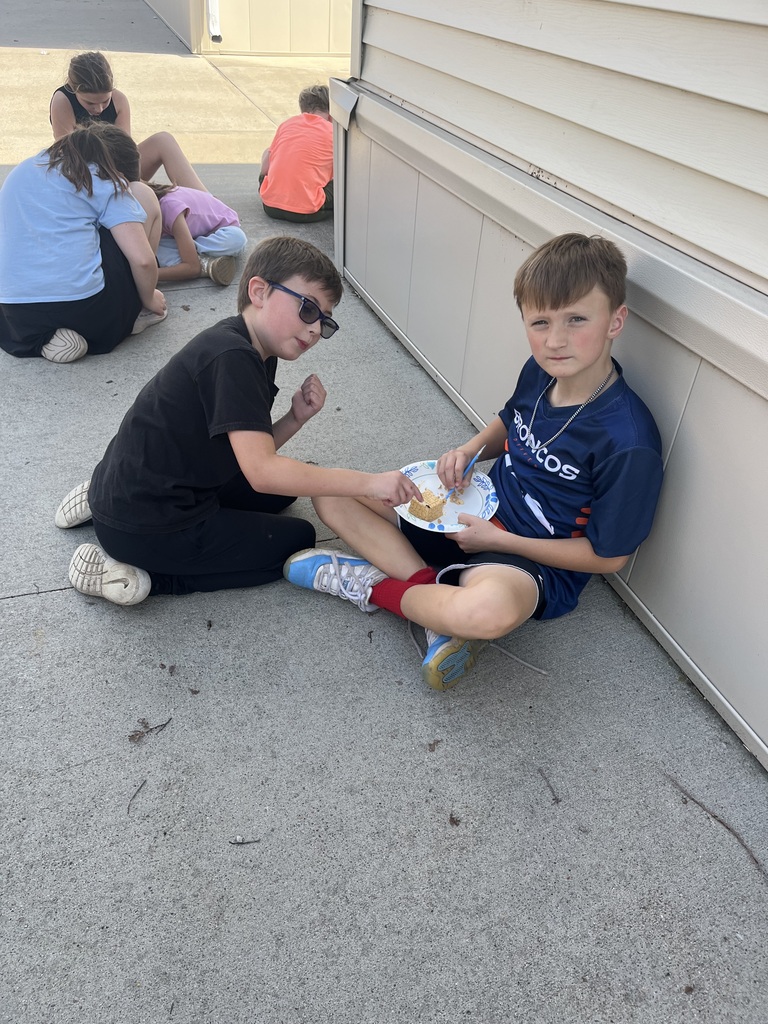 Two young boys sitting against the side of a building, using plastic knives to carefully scrape away at a small fossil block on a paper plate.