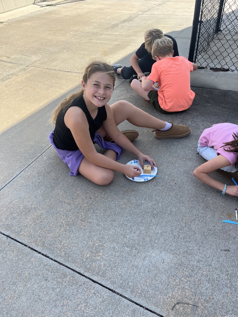 Young girl sitting cross-legged on a sidewalk smiling next to a paper plate with an excavation block during an outdoor science activity.