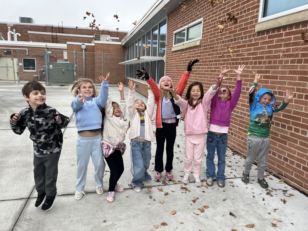 Group of young elementary students cheering with raised arms outside against a brick school building as fallen autumn leaves rain down over them.