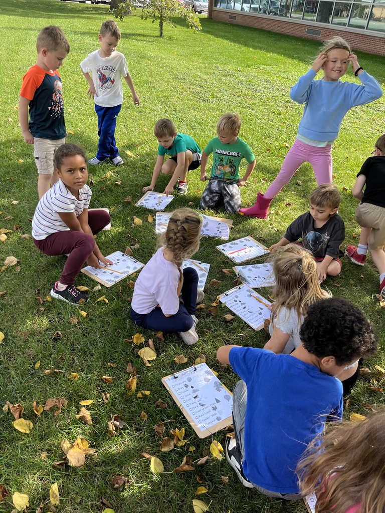 Elementary students sitting in a circle on the grass outside, reviewing their fall scavenger hunt worksheets on clipboards together.