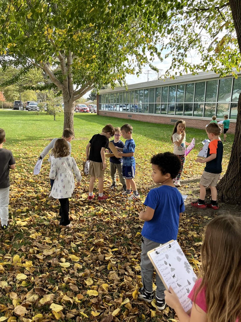 Class of young children scattered across a leaf-covered lawn outside a school building, comparing their scavenger hunt clipboards.