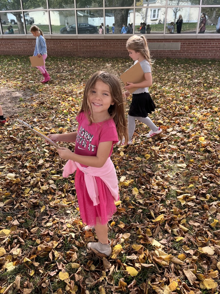 A smiling young girl in a pink shirt standing in a field covered with fallen autumn leaves while holding a clipboard.