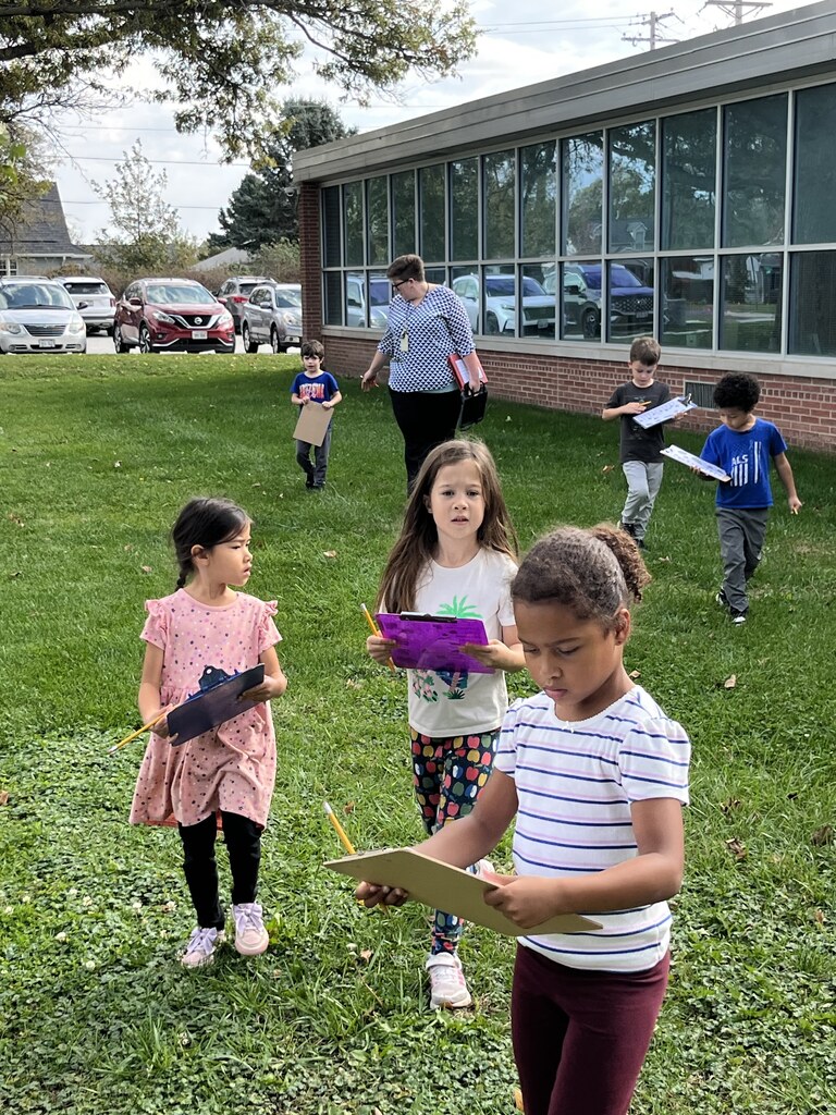 Elementary students walking through a grassy school yard holding clipboards and pencils during an outdoor fall scavenger hunt activity.