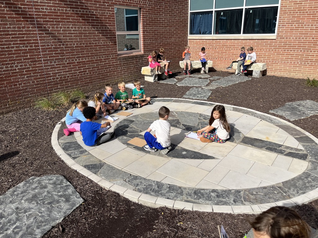 Students sitting spaced out in a decorative stone patio courtyard outside a brick school building, working on clipboards.