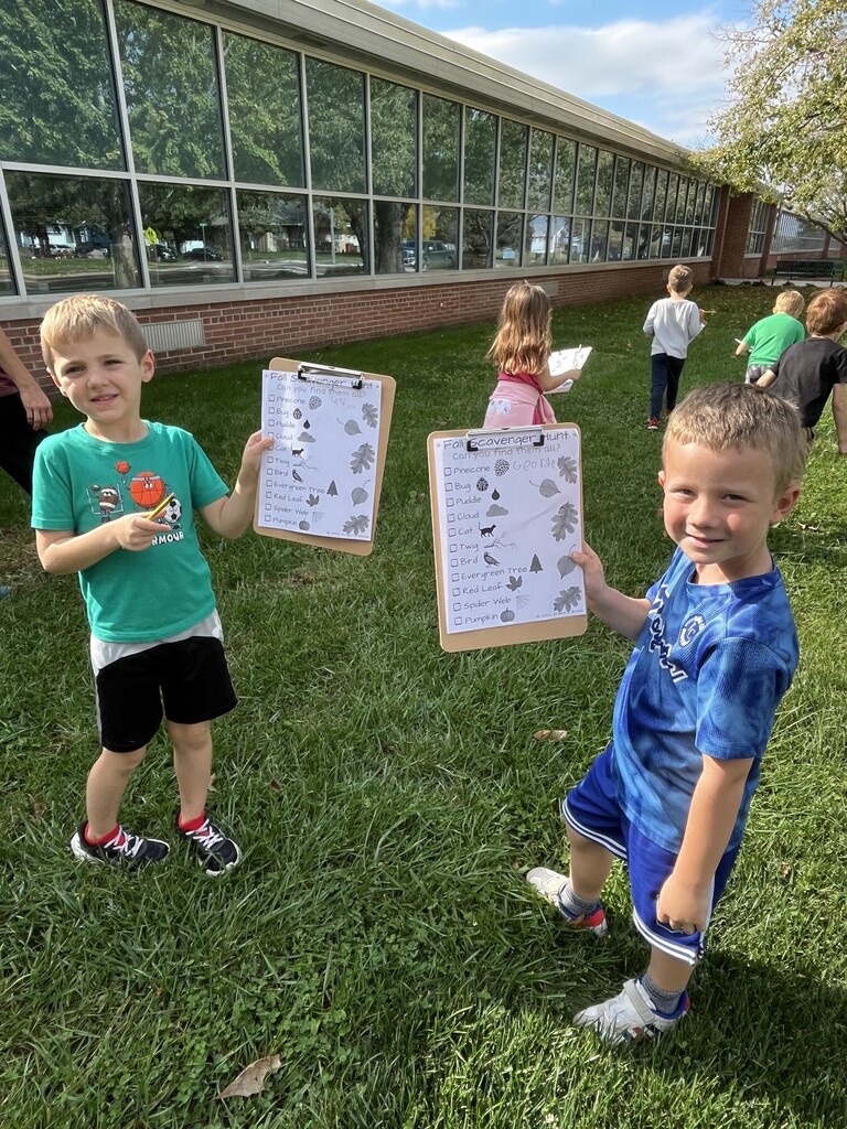 Two young boys standing on a school lawn, proudly displaying their completed fall scavenger hunt clipboards to the camera.