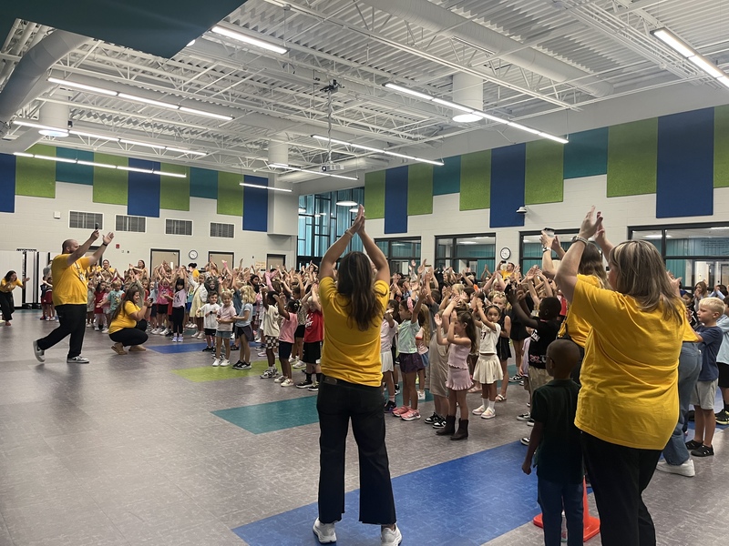 Students standing in the commons area of the school with teachers in gold shirts all around the outside. Students and staff with hands in the air clapping along to a song.